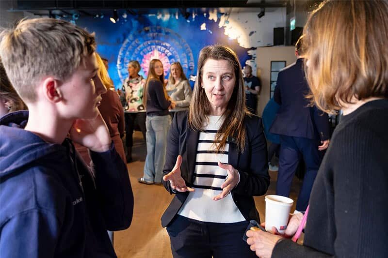 Woman speaking with two people in a casual indoor event space, with others networking in the background.