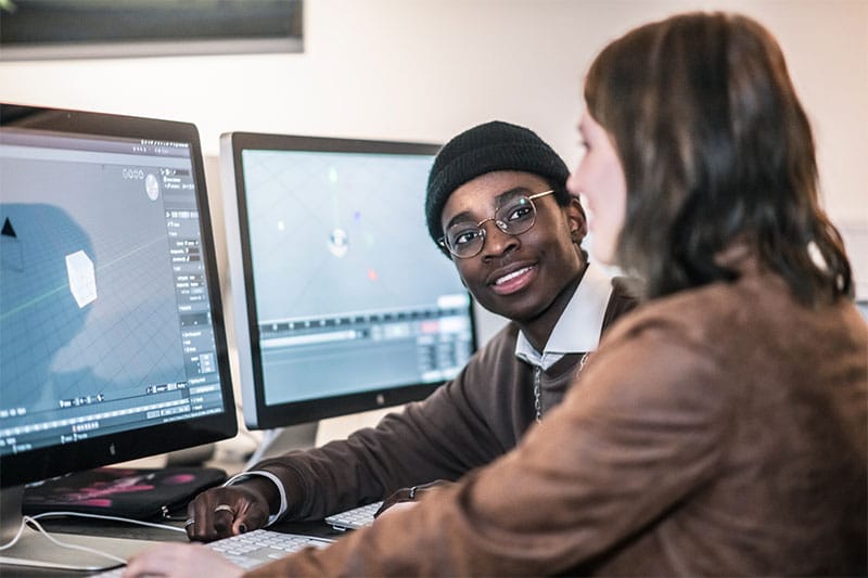 Two students working on 3D design software on desktop computers in a classroom