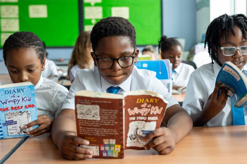 Primary school children in uniform reading 'Diary of a Wimpy Kid' books at their desks in a classroom.