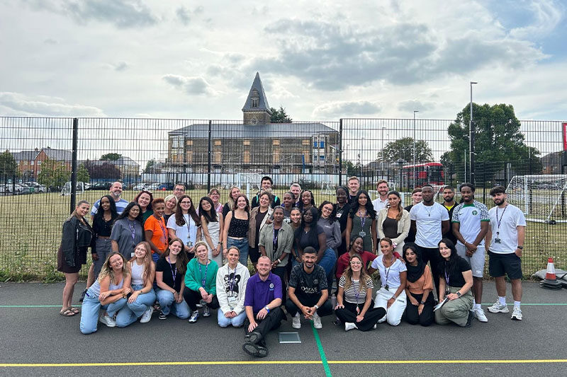 Large group of young adults and staff posing for a photo on a school playground, with a historic building in the background.