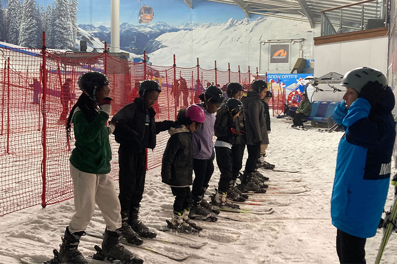 A group of students in ski gear standing in a line indoors, receiving instruction at an indoor snow centre.