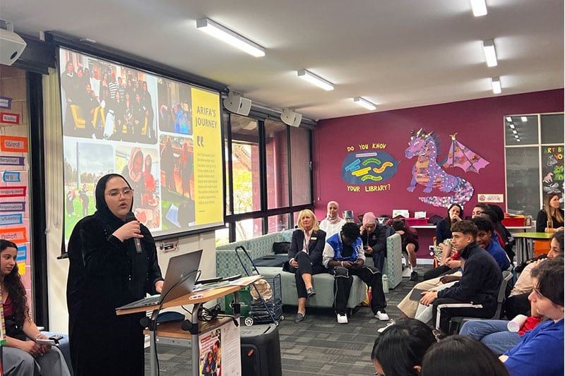 Student presenting at a school event with an audience and a slideshow titled 