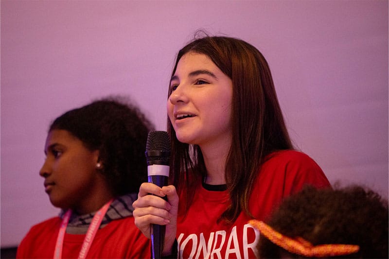 Student speaking confidently into a microphone during a school or youth event, wearing a red shirt and engaging with the audience.