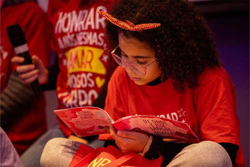 A young girl wearing glasses and a red bow headband deeply focused on reading a book, surrounded by classmates in matching red T-shirts.