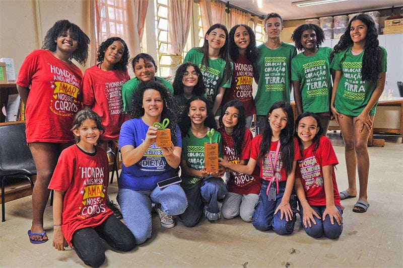 A group of smiling students and a teacher posing indoors with colourful T-shirts and wooden awards, celebrating an achievement in a classroom setting.