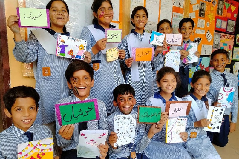 Smiling school children in uniform holding handmade Eid cards, celebrating in a colourful classroom decorated with artwork.