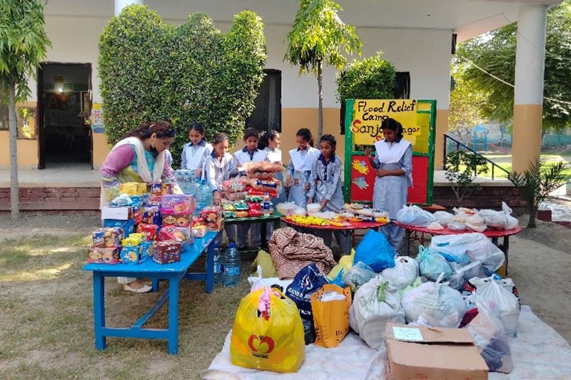 Students and a teacher organising food and supplies at a school flood relief donation camp, with tables displaying essential items and donation bags.