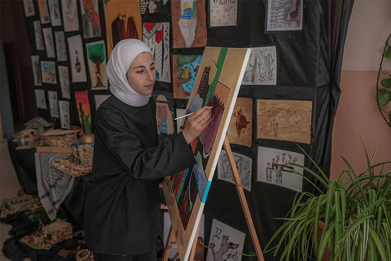 A student wearing a hijab painting on a canvas in an art exhibition space, surrounded by colourful artwork and creative displays.