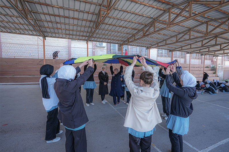 Students participating in a group activity under a covered playground, holding up a colourful parachute in a circle.