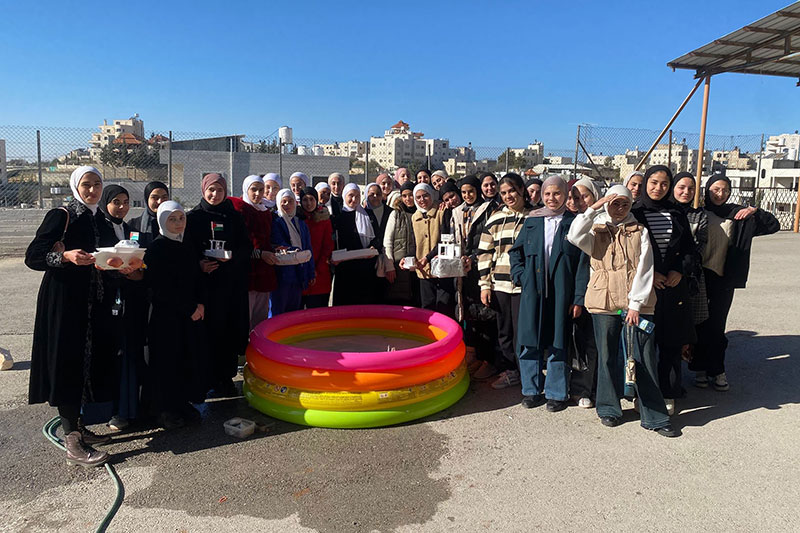 A group of students and staff posing outdoors around a colourful inflatable pool, holding model projects on a sunny day at school.
