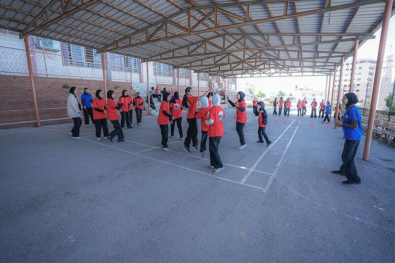 Girls in red sports uniforms playing a team game under a covered outdoor court at school, with coaches and other students observing.