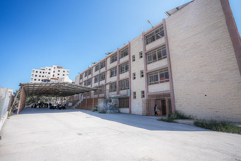 Exterior view of a multi-storey school building with a shaded outdoor area and students gathered underneath.