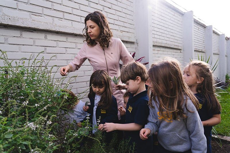 A teacher showing a group of young school children different plants in a garden by a white brick wall.