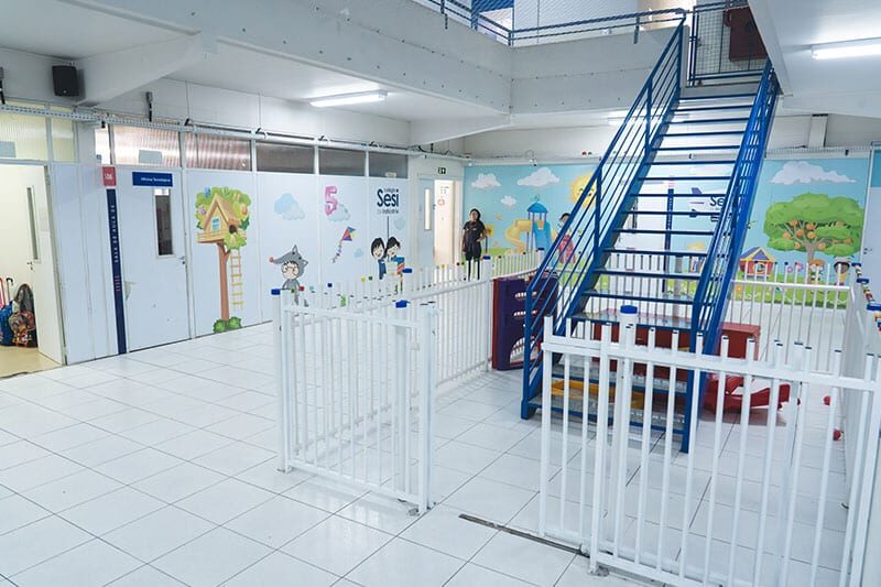 Indoor play area in a brightly lit preschool or daycare facility, featuring colorful wall murals, a blue staircase, and fenced play zones.