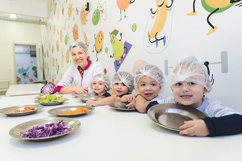 Smiling preschool children wearing hairnets sit at a table with metal plates, accompanied by a teacher, surrounded by trays of fresh vegetables in a colorful room with food-themed wall art.