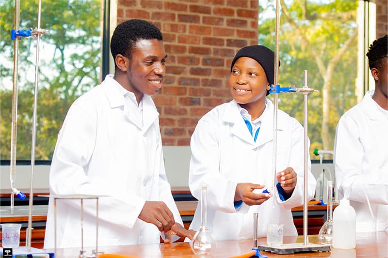 Two students in white lab coats smiling and working together on a science experiment in a laboratory.