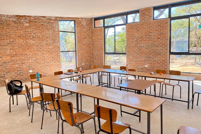 Sunlit classroom with wooden chairs and tables arranged in a U-shape, exposed brick walls, and large windows.