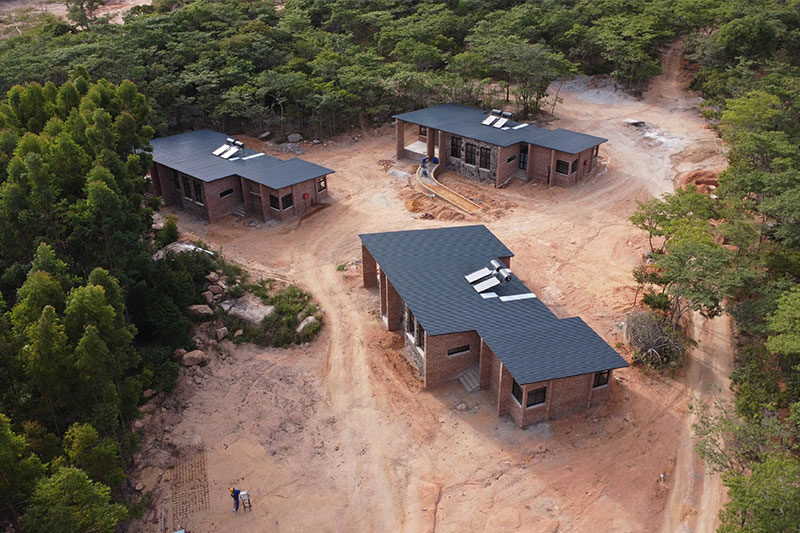 Aerial view of several modern school buildings with dark roofs and brick walls, surrounded by trees and dirt paths.