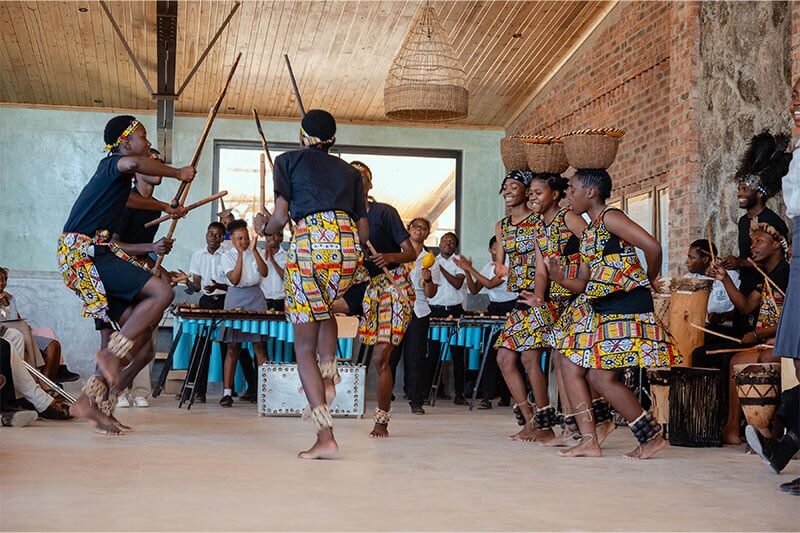 Group of young performers in colourful traditional attire dancing and playing instruments in a cultural celebration indoors.