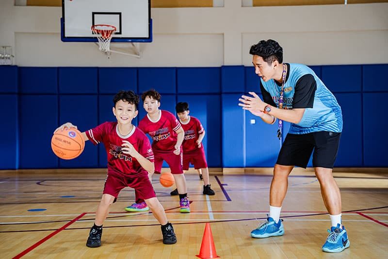 Young students in red uniforms practice basketball in a modern indoor gym with their coach.