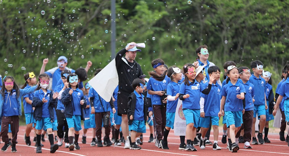Group of young students in blue uniforms and colorful masks walking on a track with teachers, surrounded by floating bubbles