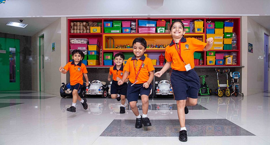 Four young children in orange school uniforms running happily inside a brightly coloured play area, with toys and storage bins neatly arranged on shelves in the background.