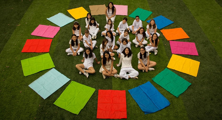 Group of students dressed in white sitting in a circle on grass, surrounded by colorful rectangular cloths arranged in a ring