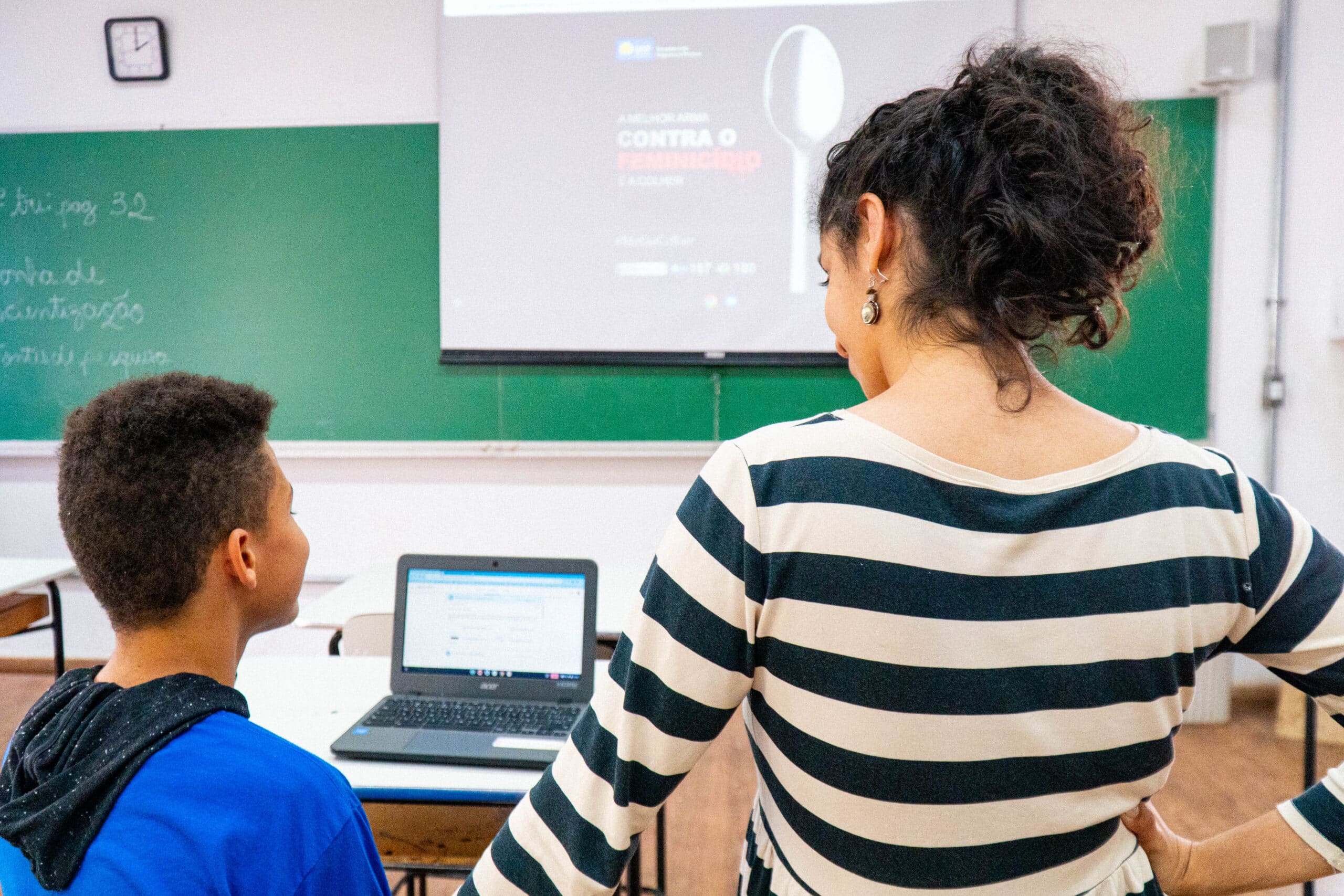 Teacher and student looking at a laptop screen in a classroom with a green chalkboard and digital projector display