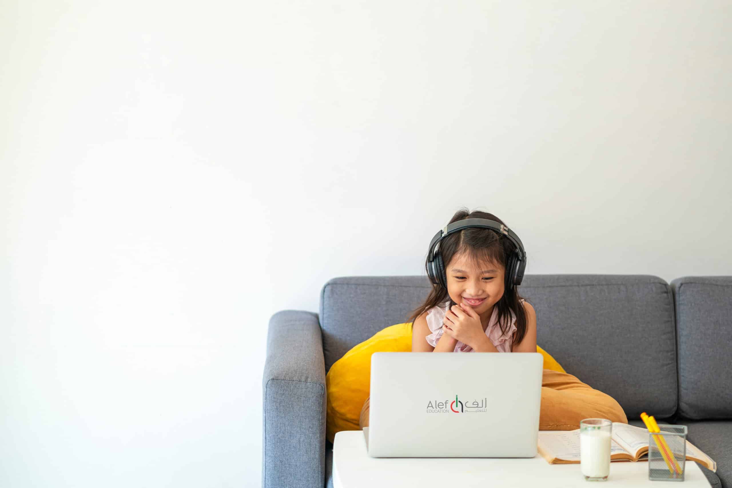 Young girl wearing headphones using a laptop at home, with a cup of milk and a notebook on the table.