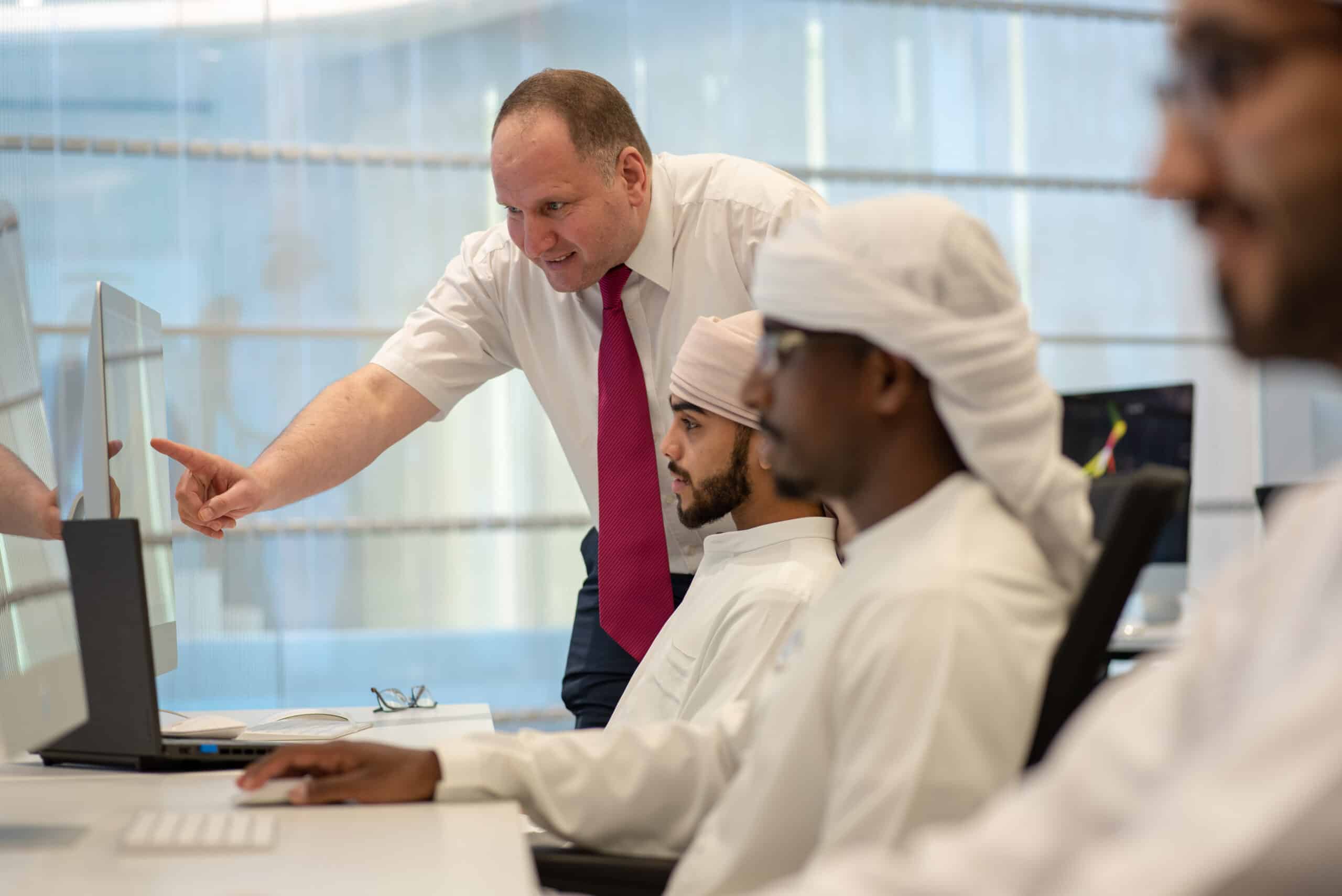 Instructor guiding a group of men in traditional attire at computer workstations in a modern training facility