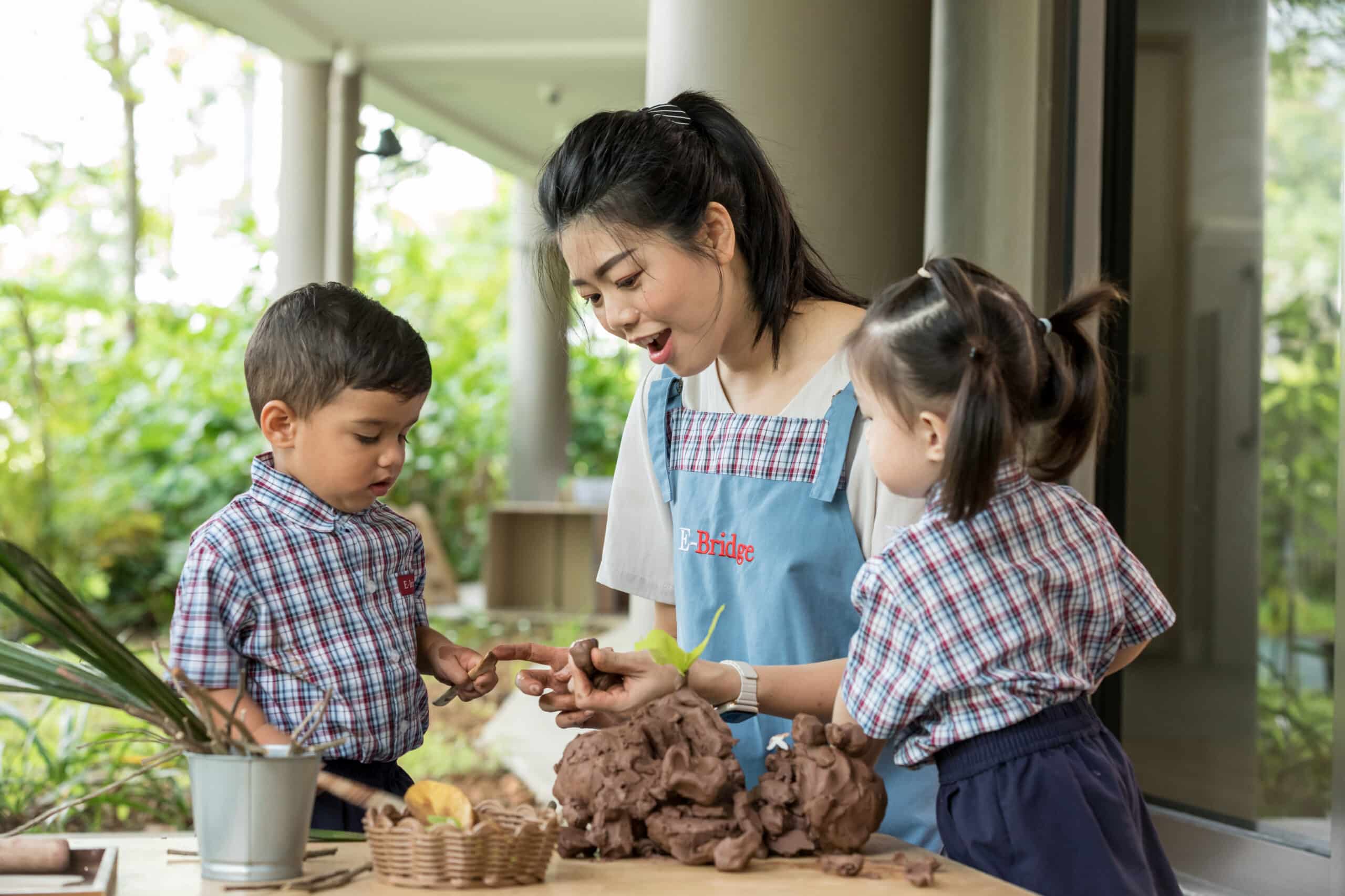 Teacher guiding two young children in a hands-on clay activity at an outdoor classroom table