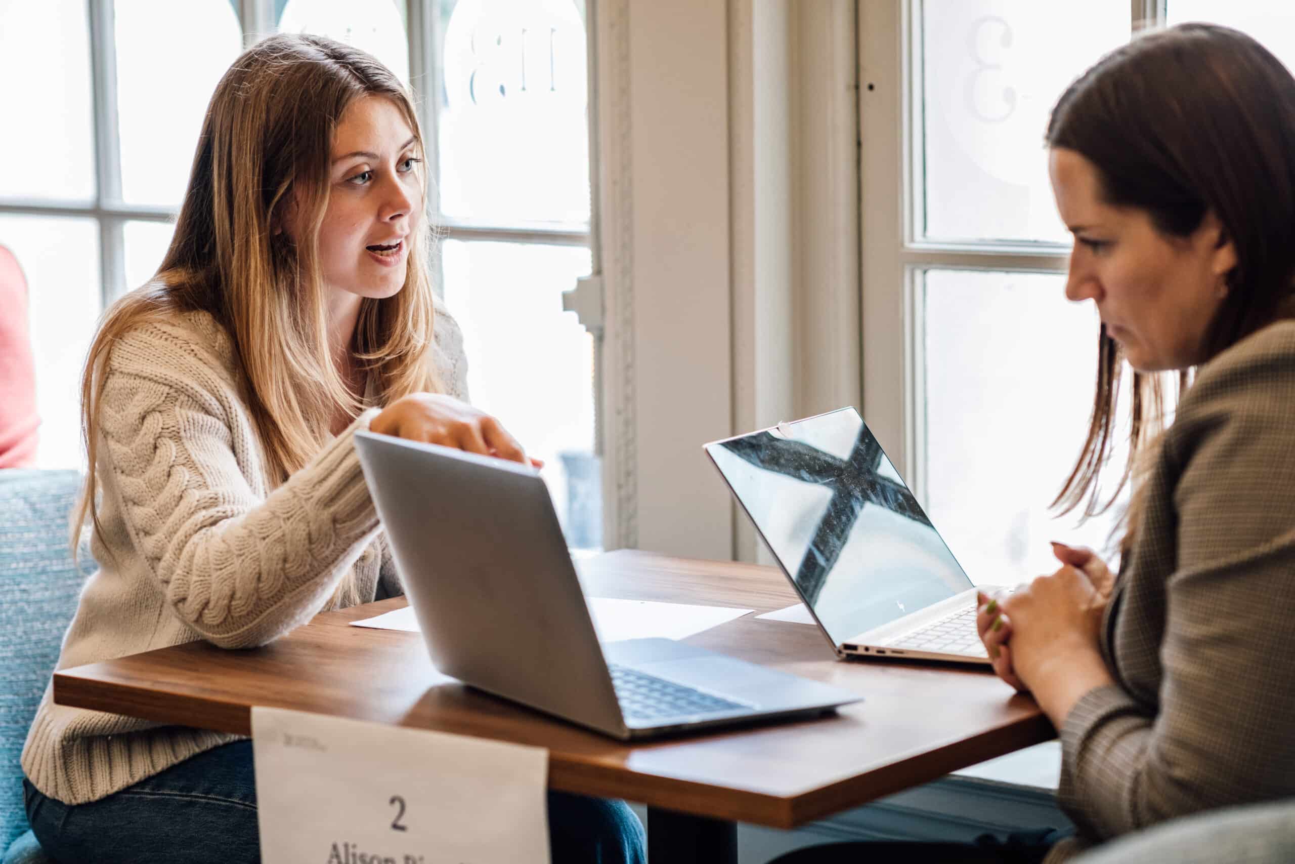 Two women discussing work or ideas at a coffee shop with laptops open on the table.