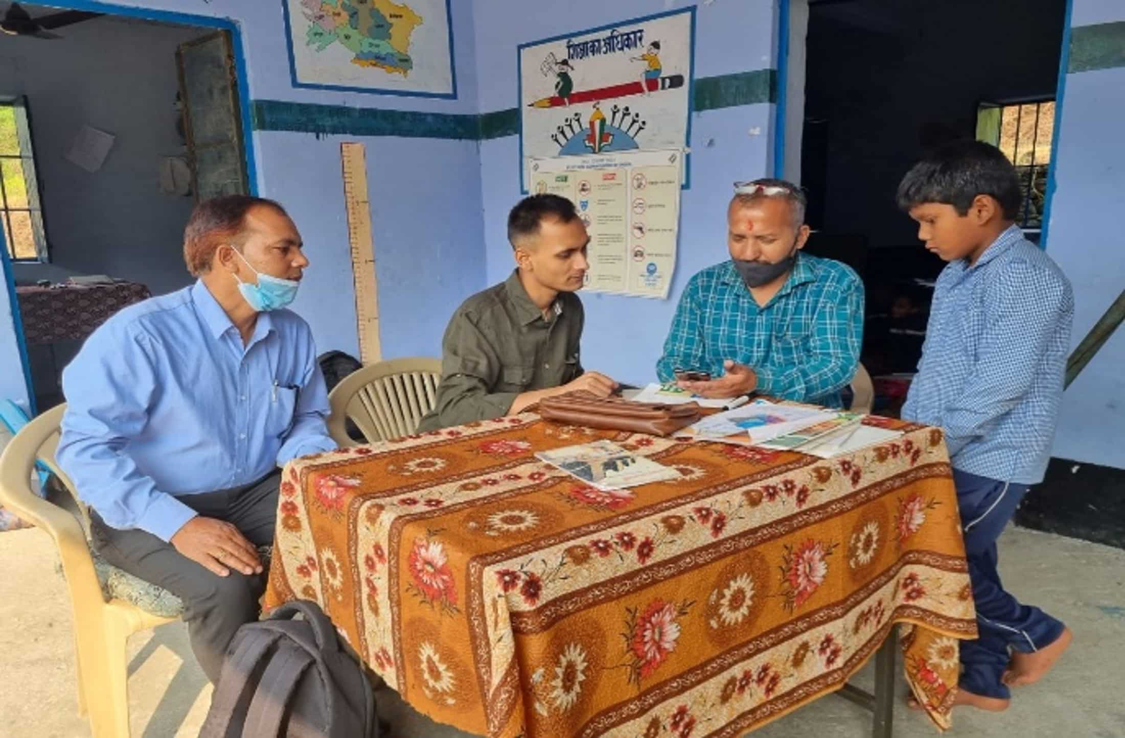 Community meeting with teachers and a guardian discussing a student’s progress at a rural school in front of educational posters
