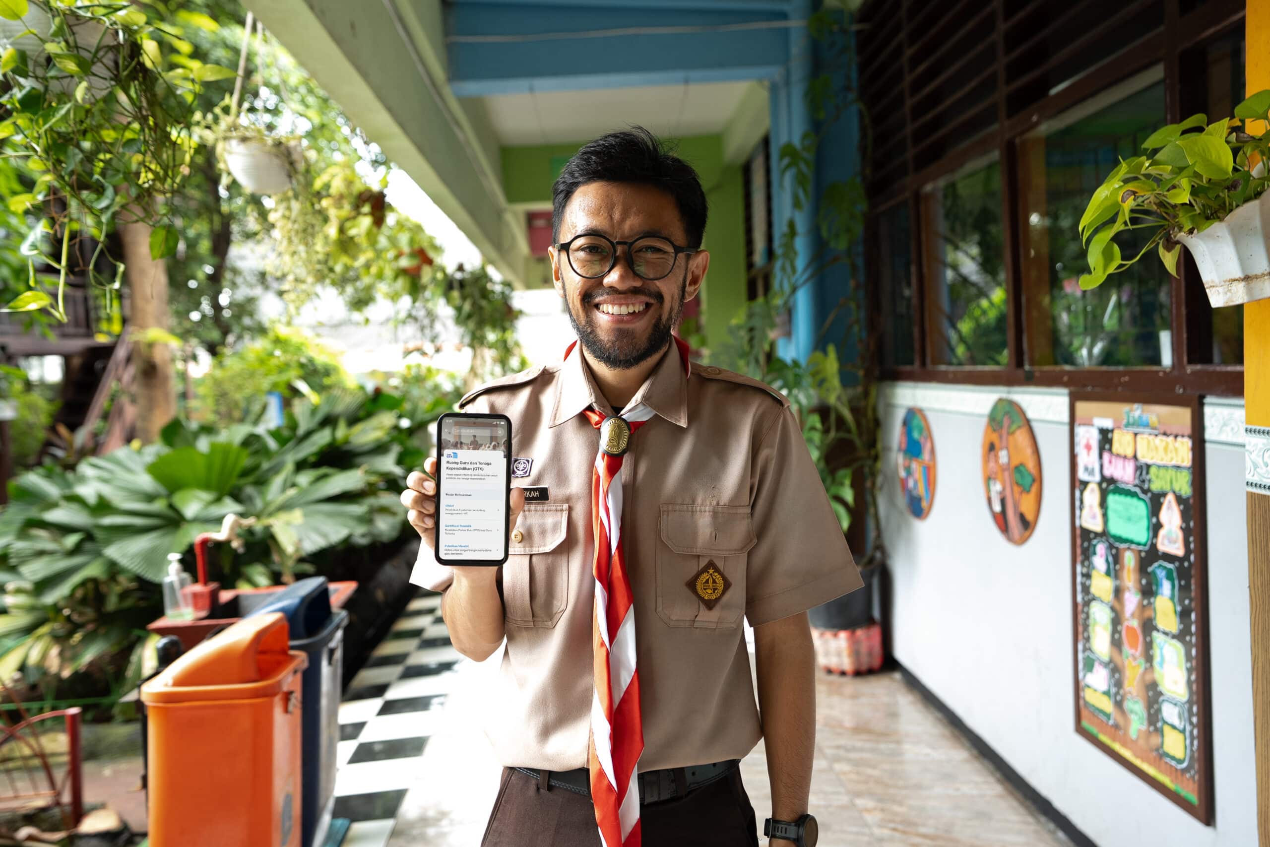 Smiling teacher in uniform holding a smartphone with educational content displayed, standing in a school corridor decorated with plants and student artwork