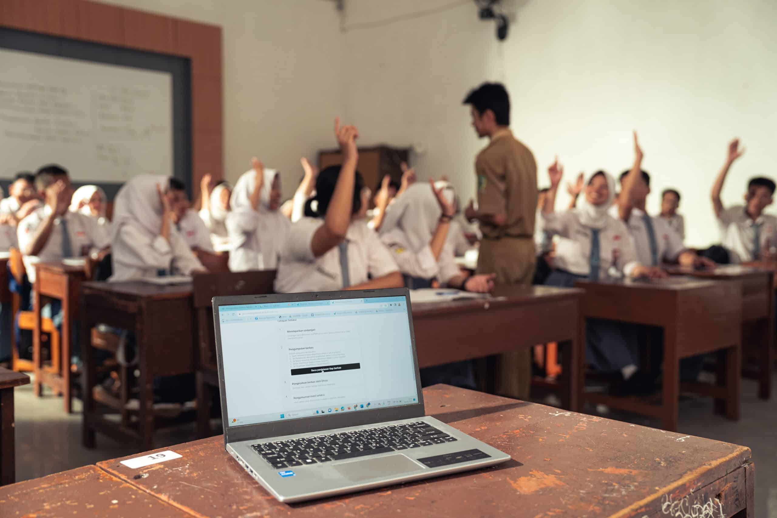 Laptop on teacher’s desk in a classroom with students raising hands and a teacher engaging with them