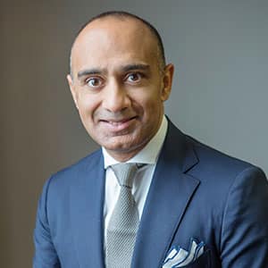 Portrait of a professionally dressed man wearing a navy blue suit, white shirt, and light gray tie, smiling at the camera.