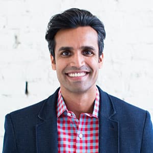 Smiling man wearing a dark blazer over a red and blue checkered shirt, standing in front of a white brick wall.