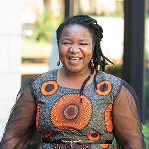 Smiling woman wearing a colorful patterned dress with circular orange designs, standing outdoors.