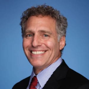 Smiling man with curly gray hair wearing a dark suit, striped shirt, and red tie against a blue background.