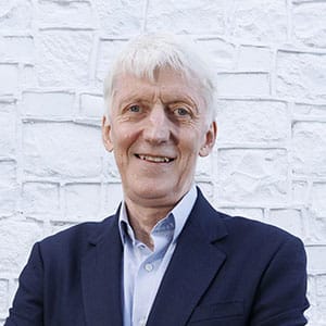 Smiling man with short white hair wearing a dark blazer and light blue shirt, standing in front of a white brick wall.