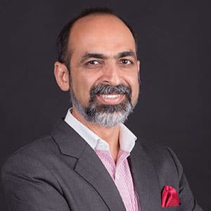 Smiling man with a beard wearing a dark suit jacket, white shirt, and pink pocket square, standing against a dark background.