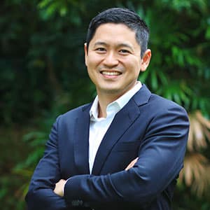 Smiling man with short dark hair wearing a dark suit and white shirt, standing outdoors with green foliage in the background.
