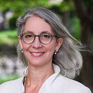 Smiling woman with gray hair wearing glasses, pearl earrings, and a light-colored top, standing outdoors.