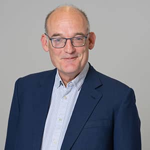 Smiling man wearing glasses, a blue blazer, and a light striped shirt, standing against a plain background.