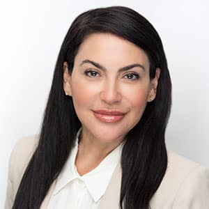 Smiling woman with long dark hair wearing a light-colored blazer and white blouse, standing against a white background.