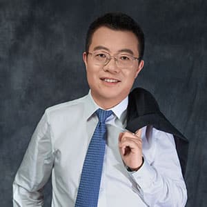 Smiling man wearing glasses, a white shirt, and a blue tie, holding a dark jacket over his shoulder against a dark backdrop.
