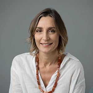 Portrait of a smiling woman with shoulder-length hair wearing a white blouse and a chunky amber necklace