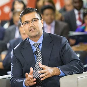 Man wearing glasses, a dark suit, and a patterned tie speaking while seated in front of an audience.