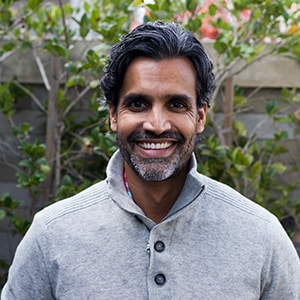 A man with dark, curly hair and a beard, smiling while wearing a light gray sweater, standing outdoors with greenery in the background.