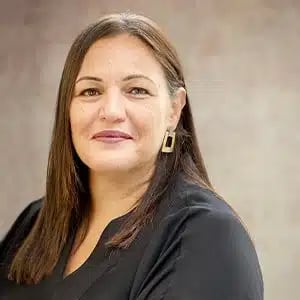A professional portrait of a woman with dark brown hair, wearing a black blouse and gold geometric earrings, with a neutral background.